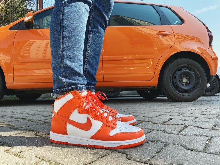 Person in blauen Jeans und roten Nike-Sneakers neben orangefarbenem Auto