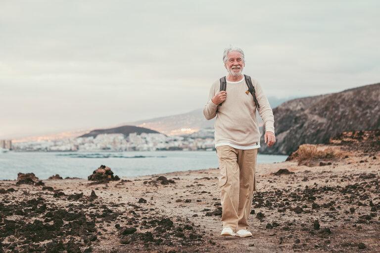 Alter älterer Mann, der auf der Klippe spazieren geht, einen Rucksack trägt und lächelt, während er einen Ausflug zum Meer im Licht des Sonnenuntergangs genießt. Aktiver weißhaariger Mann im Ruhestand