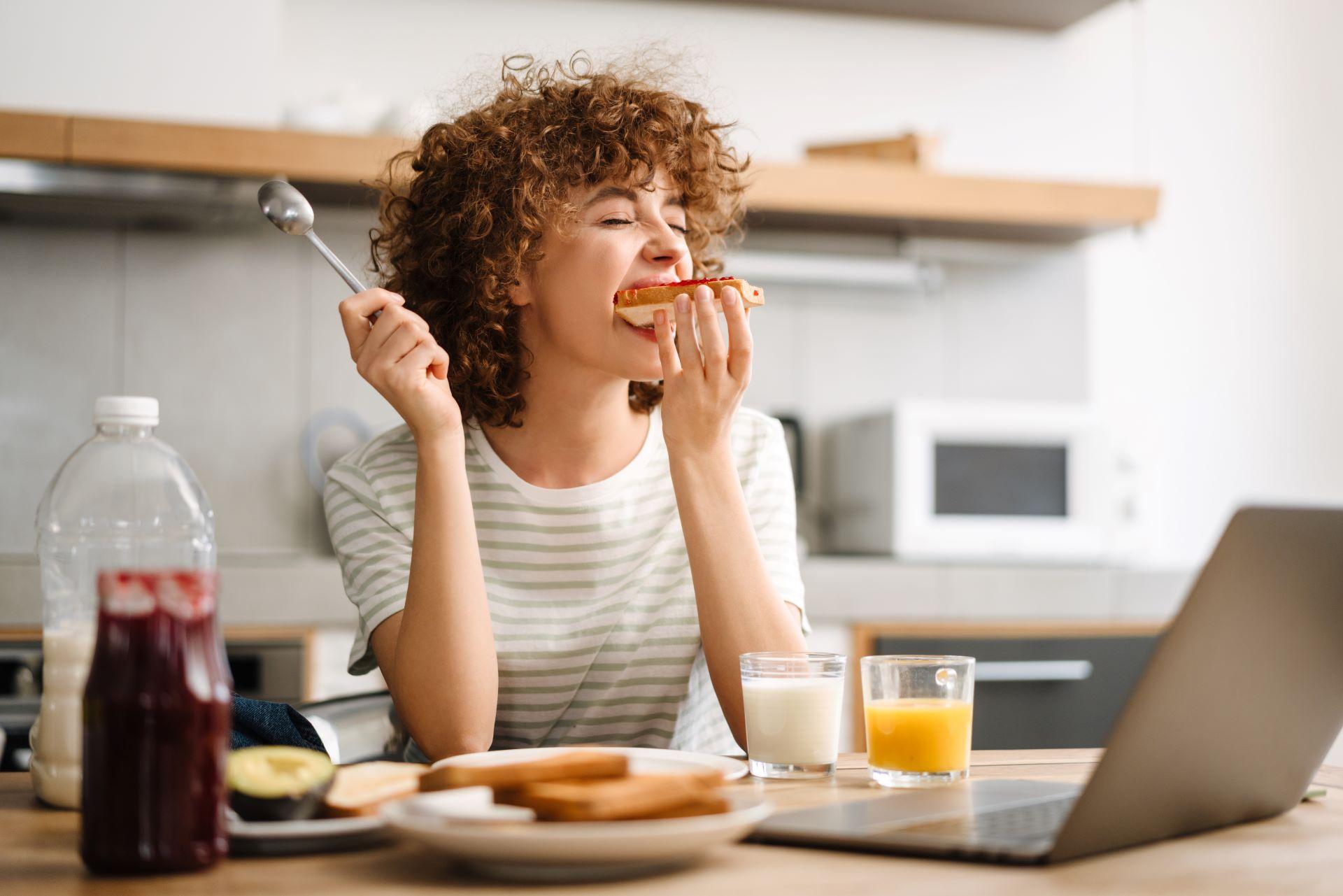 Lächelnde junge Frau, die einen Laptop benutzt, während sie zu Hause in der Küche frühstückt