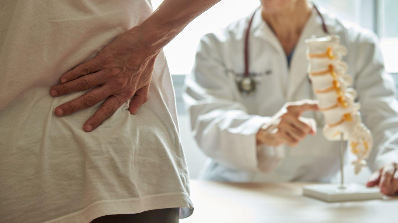 Patient mit Rueckenschmerzen zeigt auf die Huefte, waehrend Orthopäde Köln Wirbelsaeulenmodell zur Erklaerung nutzt